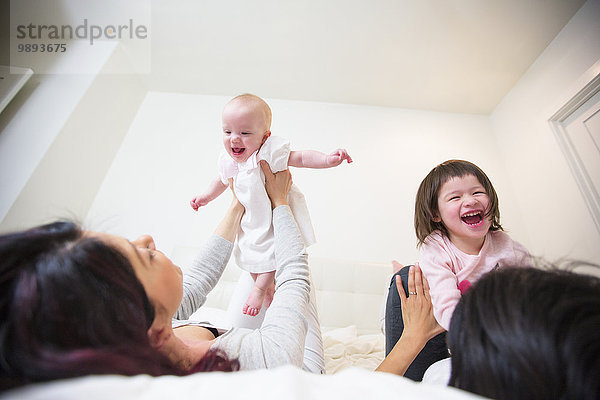 Über die Schulteransicht eines weiblichen Paares  das mit Baby- und Kleinkindtöchtern auf dem Bett lacht.