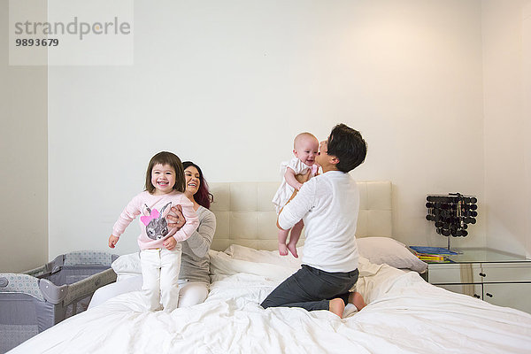 Weibliches Paar auf dem Bett  das mit Baby- und Kleinkindtöchtern spielt.