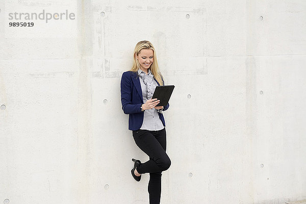 Lächelnde Geschäftsfrau mit digitalem Tablett an Betonwand gelehnt
