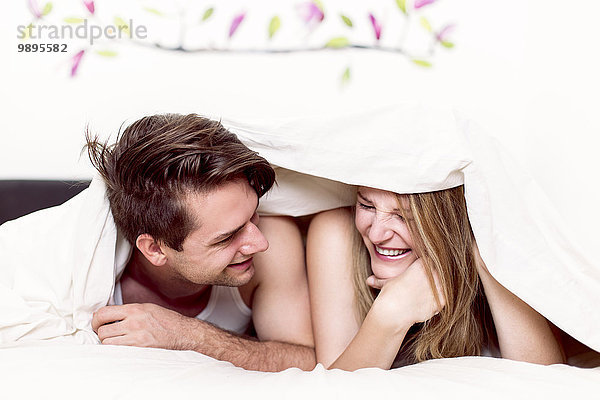 Lachendes Paar liegt nebeneinander im Bett unter der Decke.