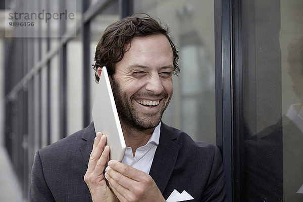 Porträt eines lachenden Geschäftsmannes  der versucht  mit seinem digitalen Tablett zu telefonieren.