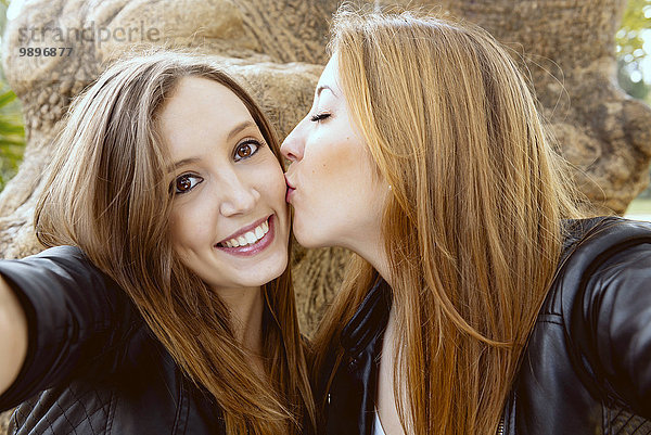 Junge Frau nimmt einen Selfie  während ihre Freundin sie auf die Wange küsst.
