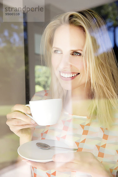 Blonde Frau steht am Fenster und trinkt Kaffee.