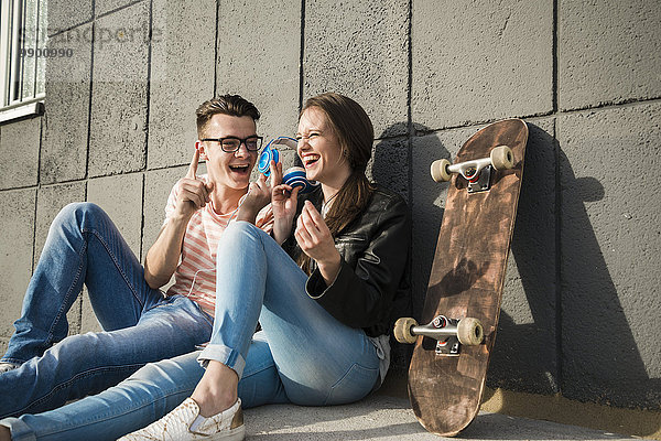 Glückliches junges Paar auf dem Boden sitzend mit Kopfhörer und Skateboard