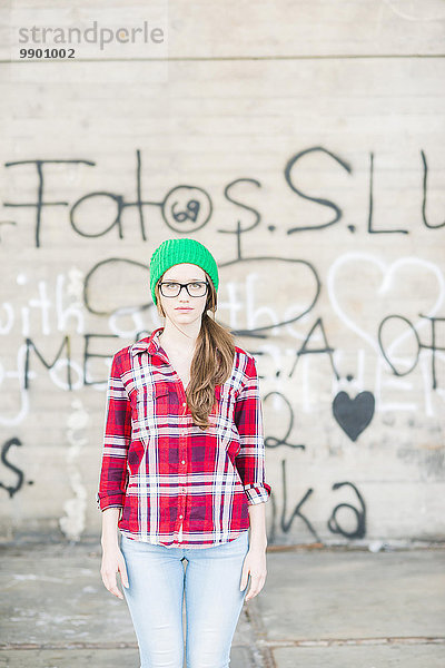 Junge Frau mit kariertem Hemd und grünem Wollhut an der Graffiti-Wand