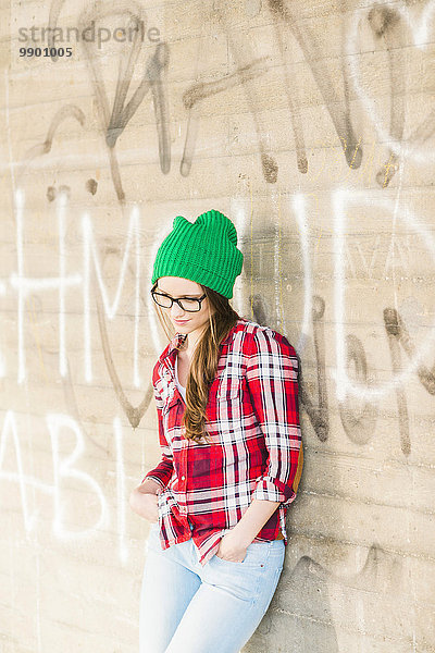 Junge Frau mit kariertem Hemd und grünem Wollhut an der Graffiti-Wand lehnend