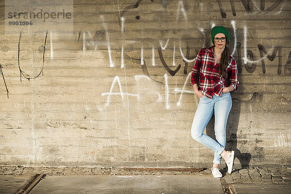 Junge Frau mit kariertem Hemd und Wollmütze an Graffiti-Wand lehnend