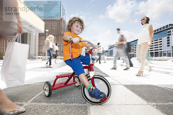Ein Junge auf einem Dreirad zwischen einer Menschenmenge in einer Stadt.