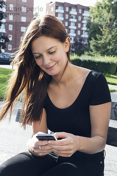 Lächelnde junge Frau mit Smartphone auf der Bank im Freien