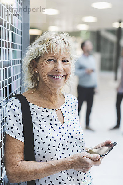 Porträt einer glücklichen Geschäftsfrau  die ein digitales Tablett hält  während sie sich im Büro an die Wand lehnt.