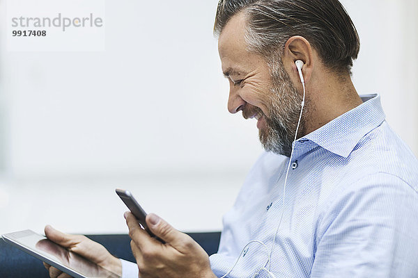 Glücklicher Geschäftsmann mit digitalem Tablett beim Musikhören per Handy in der Büro-Lobby