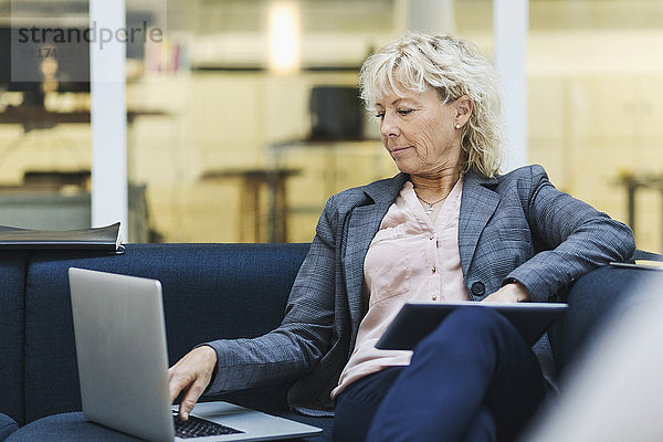 Geschäftsfrau mit Laptop und digitalem Tablett auf dem Sofa in der Bürolobby