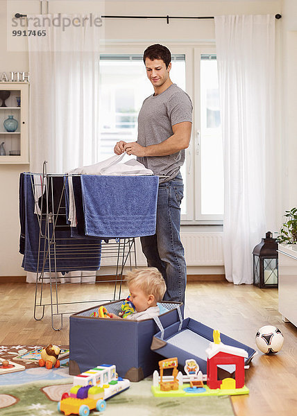 Vater trocknet Kleidung  während er den kleinen Jungen beim Spielen mit Spielzeug beobachtet.