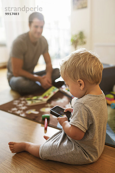 Baby Junge mit Handy auf dem Boden  während Vater mit Spielzeug im Hintergrund sitzt.