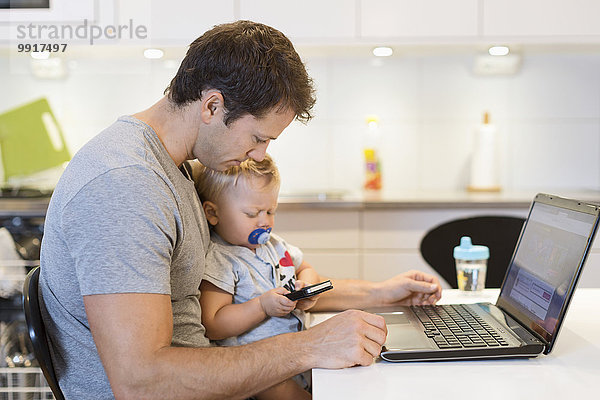 Vater mit Laptop  der den kleinen Jungen mit dem Handy am Tisch ansieht.