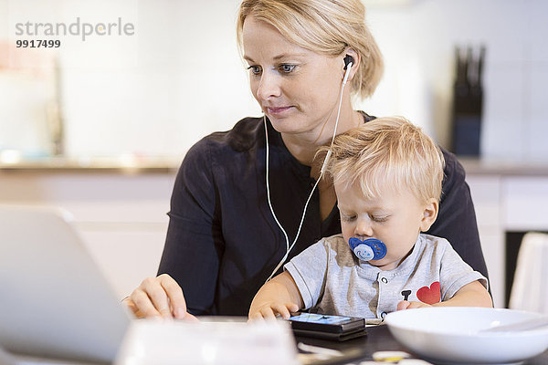Baby Junge mit Smartphone  während die Mutter Musik auf dem Laptop am Tisch hört.
