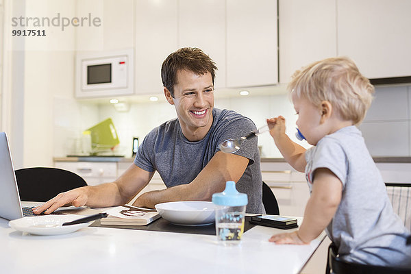 Glücklicher Vater schaut auf den kleinen Jungen  während er den Laptop in der Küche benutzt.