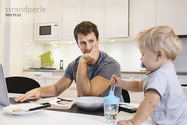 Vater schaut auf den kleinen Jungen  während er den Laptop in der Küche benutzt.