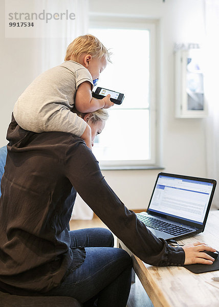Mutter mit Laptop beim Tragen des Jungen auf den Schultern zu Hause
