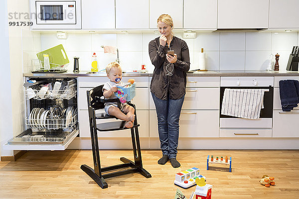 Mutter spricht auf dem Handy in der Küche  während der Junge auf dem Hochstuhl sitzt.