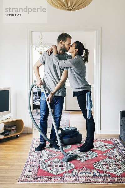 Volle Länge des Mannes  der die Frau küsst  während er den Teppich zu Hause absaugt.