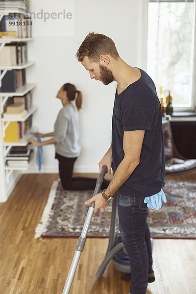 Seitenansicht des Mannes beim Staubsaugen des Bodens  während die Frau zu Hause im Hintergrund Regale putzt.