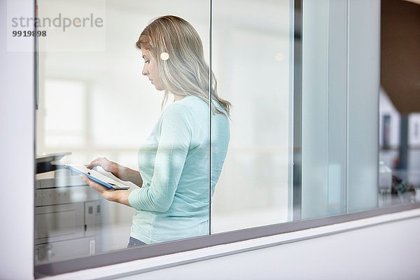 Mittlere erwachsene Geschäftsfrau mit digitalem Tablett hinter dem Bürofenster