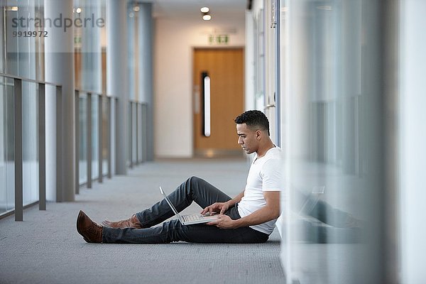 Junger Geschäftsmann sitzt im Büroflur mit Laptop