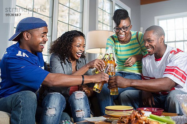 Vier erwachsene Freunde stoßen mit Flaschenbier auf dem Wohnzimmersofa an.