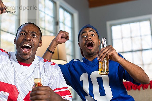 Zwei männliche Freunde feiern  während sie vom Sofa aus fernsehen.