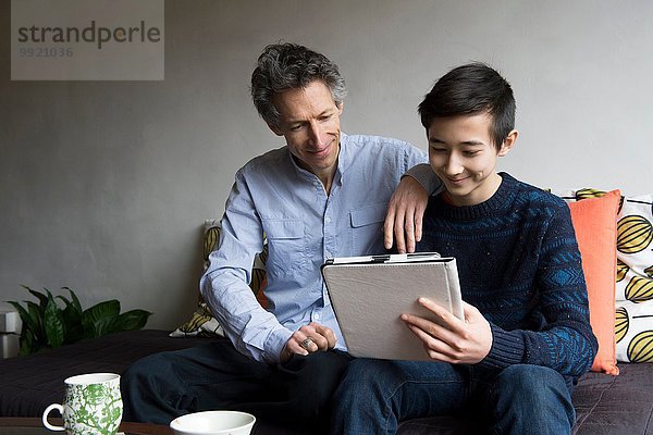 Vater und jugendlicher Sohn lesen digitales Tablett auf Sofa