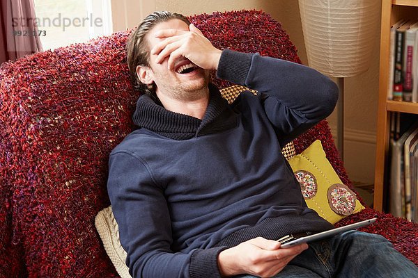 Junger Mann liegt auf dem Sofa und bedeckt sein Gesicht  während er lacht.