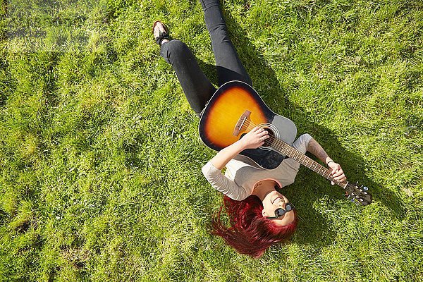 Draufsicht auf eine junge Frau  die auf Gras liegt und Akustikgitarre spielt.