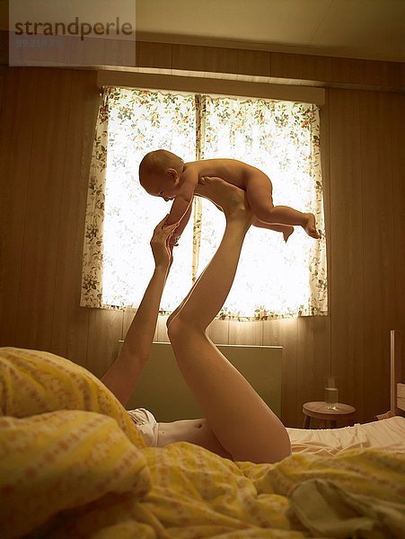 Mutter liegt auf dem Bett und balanciert den Sohn auf den Füßen.