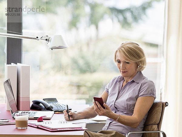 Reife Geschäftsfrau beim Lesen von Textnachrichten auf dem Smartphone am heimischen Schreibtisch