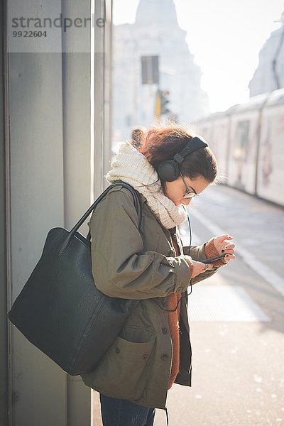 Junge Frau mit Kopfhörer beim Lesen von Smartphone-Texten auf der Straße