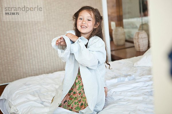 Portrait eines jungen Mädchens  das auf dem Bett sitzt und ein übergroßes weißes Hemd trägt.