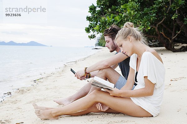 Junges Paar sitzt am Strand und liest Buch und SMS  Kradan  Thailand