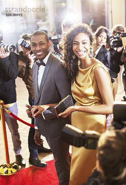 Lächelndes Promi-Paar wird von Paparazzi-Fotografen auf dem roten Teppich fotografiert.
