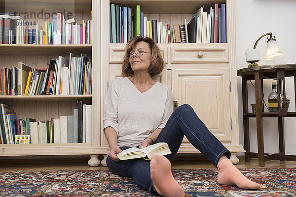 Senior woman sitting on the floor in front of bookshelf and reading  Munich  Bavaria  Germany