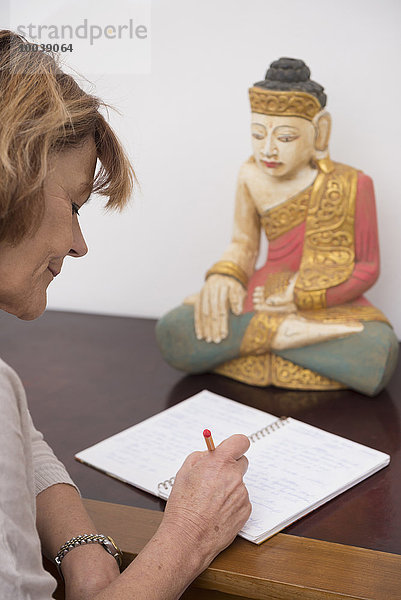 Senior woman writing in notebook  Buddha statue in the background  Munich  Bavaria  Germany