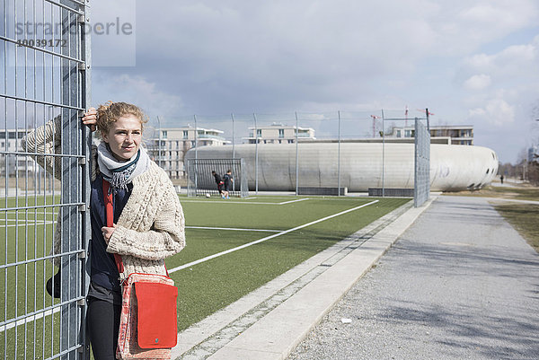 Young woman with metal fence at soccer field with players in the background  Munich  Bavaria  Germany