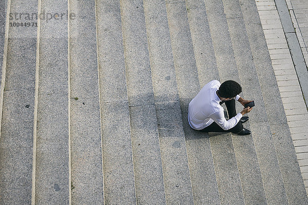 Junger Mann sitzt auf einer Treppe und schaut auf sein Handy.
