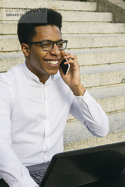 Lächelnder junger Mann auf der Treppe sitzend mit Handy und Laptop