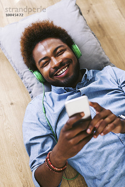 Junger Afroamerikaner auf dem Boden liegend mit Kopfhörer und Smartphone