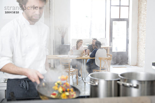 Koch arbeitet in der Küche seines Restaurants  während die Gäste im Hintergrund kommunizieren.