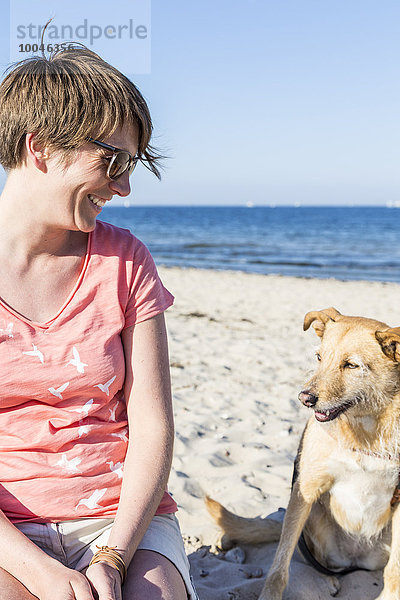 Deutschland  Kiel  lächelnde Frau sitzend mit ihrem Hund am Sandstrand