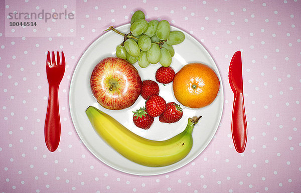 Teller mit Früchten mit lustigem Gesicht und rotem Plastikbesteck auf rosa Tuch