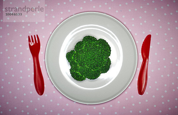 Teller mit Brokkoli und rotem Plastikbesteck auf rosa Tuch