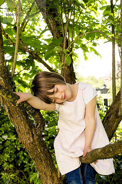 Porträt eines lächelnden Mädchens mit geschlossenen Augen auf einem Ast sitzend